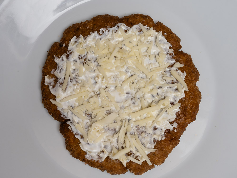 Aranybarnára sült gluténmentes lángos tejmentes tejföllel megkenve és növényi sajttal megszórva fehér tányéron tálalva.