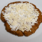 Tej- és gluténmentes lángos tejfölpótlóval megkenve, növényi sajttal megszórva, fehér tányéron tálalva - enyhén döntött szögből fotózva.