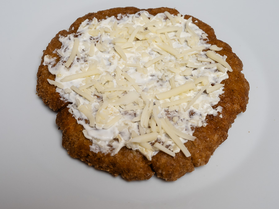 Tej- és gluténmentes lángos tejfölpótlóval megkenve, növényi sajttal megszórva, fehér tányéron tálalva - enyhén döntött szögből fotózva.