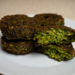 Aranybarnára sült, ropogós falafel pogácsák fehér tányéron, egy félbevágott darabbal, amely megmutatja a zöld, fűszeres belsőt.