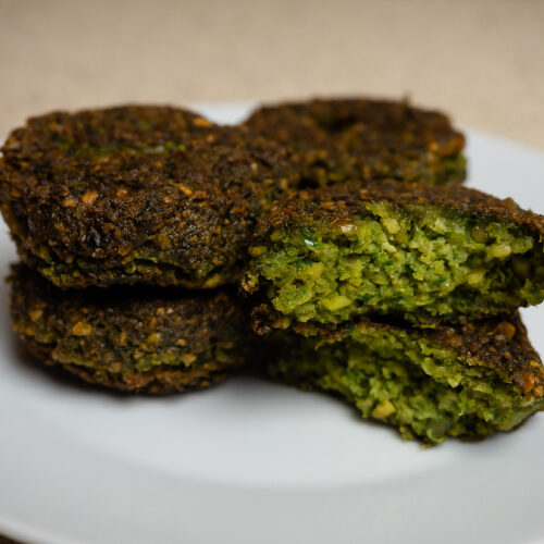 Aranybarnára sült, ropogós falafel pogácsák fehér tányéron, egy félbevágott darabbal, amely megmutatja a zöld, fűszeres belsőt.