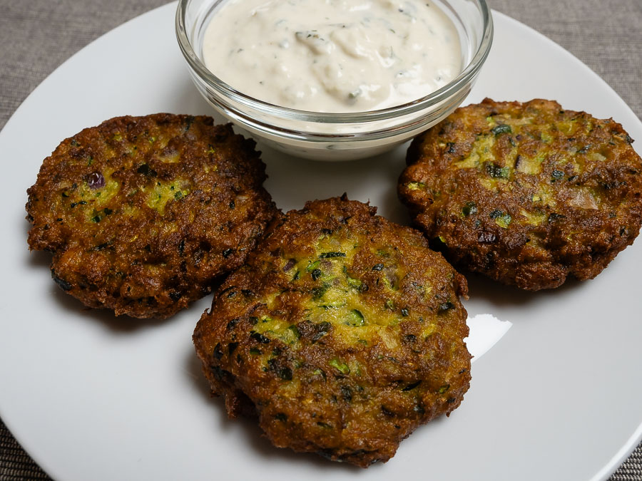 Három aranybarnára sült görög cukkini fasírt krémes vegán tzatzikivel fehér tányéron tálalva